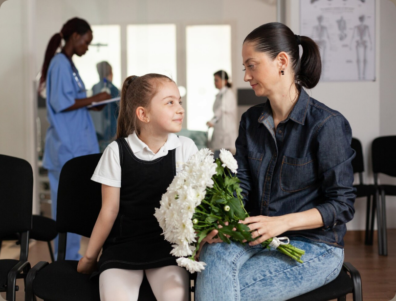 Child holding white flowers while seated with a parent in a hospital waiting area
